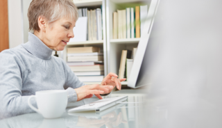 Das Bild zeigt eine ältere Frau, die mit einer Tasse Kaffee vor einem Computer sitzt.