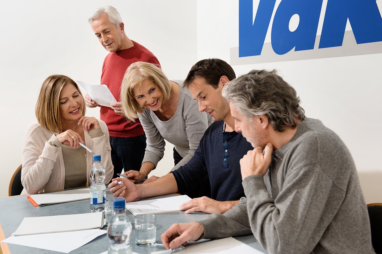 Verschiedene Menschen sitzen an einem Tisch, offenbar in einer Arbeitssitzung, an der Wand hinter ihnen ist ein gro&szlig;es VdK-Logo.