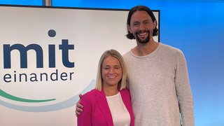 Verena Bentele und Neven Subotic stehen nebeneinander, im Hintergrund sieht man ein Display mit dem Logo der VdK-Sendung "Miteinander"