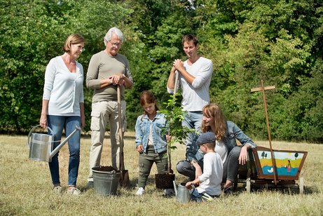 Eine Gruppe von M&auml;nnwern und Frauen ist drau&szlig;en, pflanzt einen Baum. Ein Mann h&auml;lt einen Spaten, eine Frau eine Gie&szlig;kanne. 
