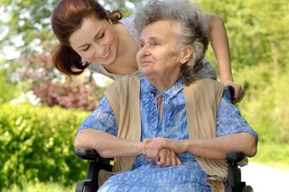 Das Bild zeigt eine ältere Frau im Rollstuhl, die von einer Frau angeschoben wird. 