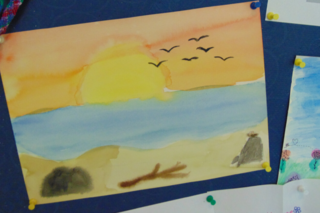 Das Foto zeigt ein mit Wasserfarben gemaltes Bild. ZU sehen sind ein Sonnenuntergang am Meer, am Strand sitzt eine Person.