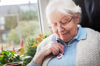 Eine Seniorin sitzt entspannt in einem Sessel und guckt auf ihr Notrufgerät, dass wie eine Kette um ihren Hals hängt.