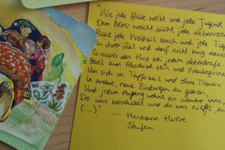 Das Bild zeigt einen gelben Zettel mit der ersten Strophe des Gedichtes "Stufen" von Hermann Hesse und daneben ein Kärtchen, das mit verschiedenen Bildchen beklebt wurde.