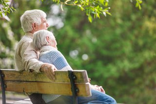 Das Bild zeigt ein älteres Ehepaar arm in arm auf einer Parkbank. 