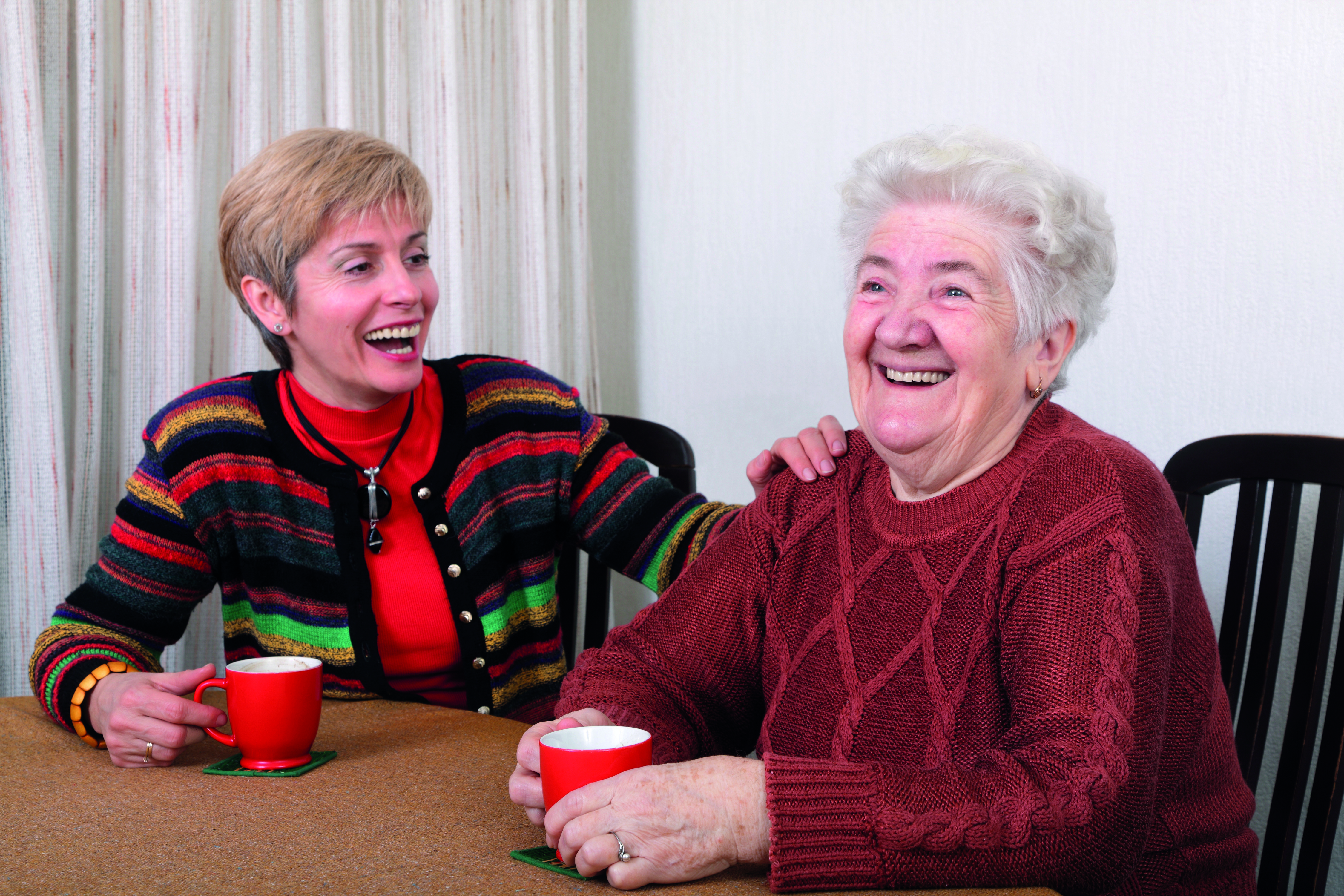 Auf dem Foto ist eine jüngere und eine ältere Frau an einem Tich zu sehen mit einer Tasse in der Hand. Die beiden freuen sich und sind am Lachen.