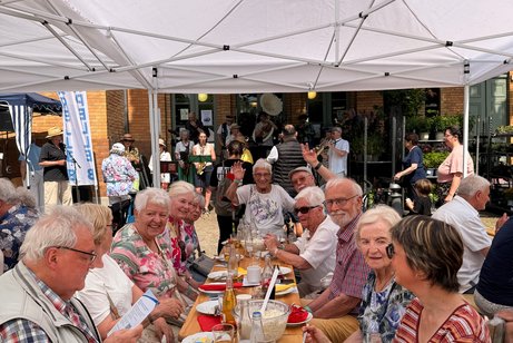 Menschen sitzen unter einem wei&szlig;en Zelt und feiern ein Sommerfest mit Essen und fr&ouml;hlicher Atmosph&auml;re.