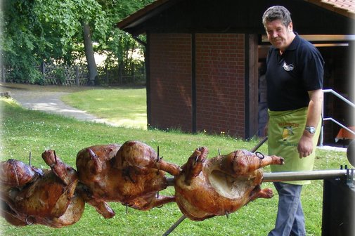 Drei ganze Brath&auml;hnchen am Spie&szlig; &uuml;ber offenem Feuer im Freien, daneben ein Mann mit Sch&uuml;rze und schwarzem Hemd