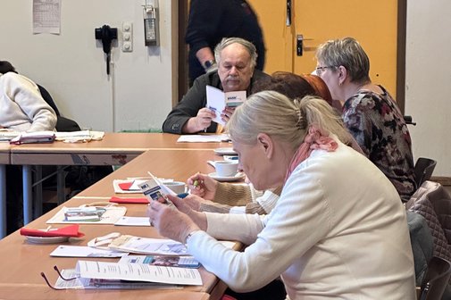 Mehrere Personen sitzen an Tischen. Eine Person l&auml;uft im Hintergrund entlang. Es lassen sich zwei Teilnehmende genauer erkennen, die in einen VdK-Flyer oder eine Brosch&uuml;re vertieft sind. Auf den Tischen liegen weiteres Infomaterial, Brillen, Geschirr und Kugelschreiber.
