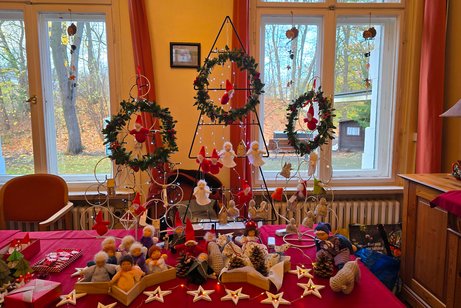 Weihnachtliche Dekoration mit handgemachten Engel- und Wichtelfiguren, Adventskränzen und Sternen auf rotem Tisch vor großen Fenstern