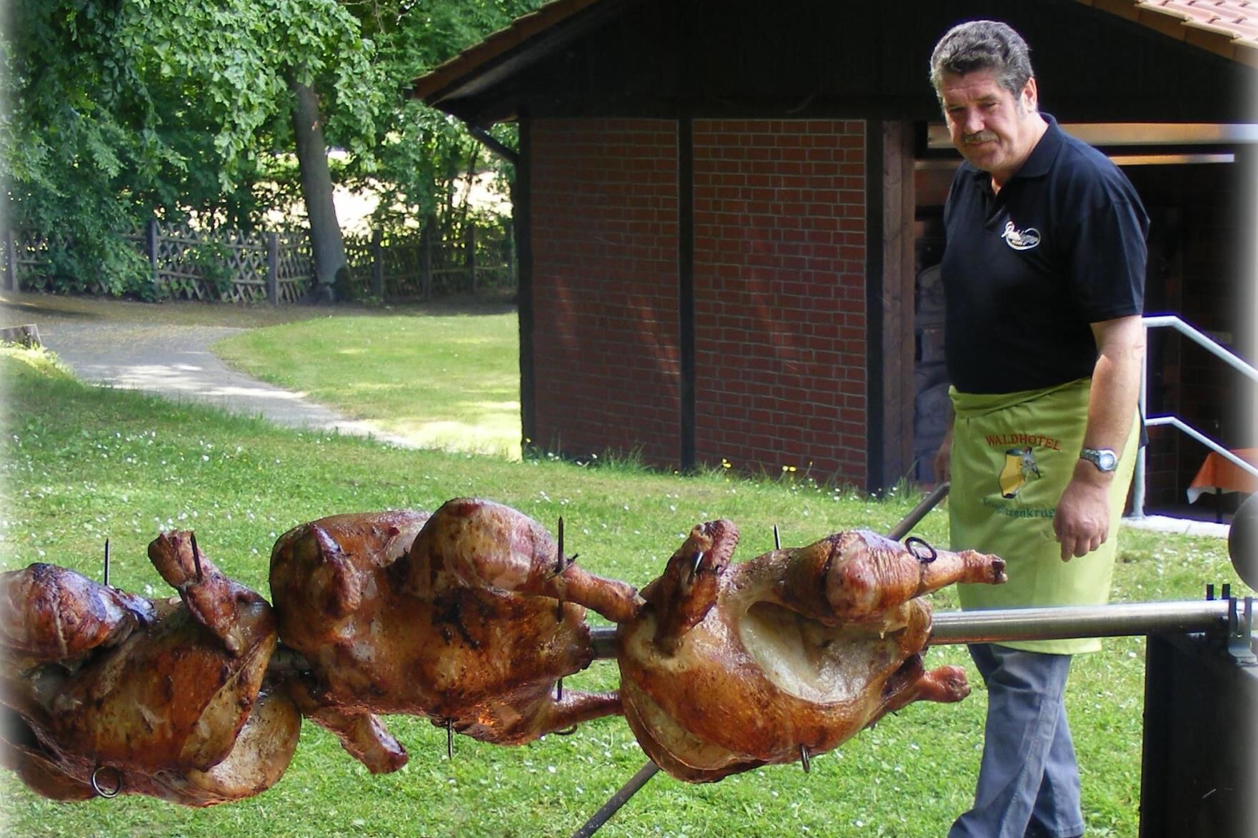 Drei ganze Brath&auml;hnchen am Spie&szlig; &uuml;ber offenem Feuer im Freien, daneben ein Mann mit Sch&uuml;rze und schwarzem Hemd